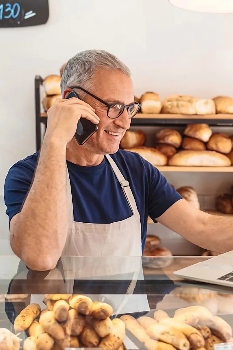 Comerciante de cabelos grisalhos atendendo ao telefone atrás do balcão da própria padaria, com o notebook aberto ao lado